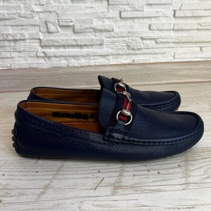 May include: A pair of navy blue leather loafers with a silver-tone metal accent and a red and navy stripe. The shoes are displayed on a light wood surface.