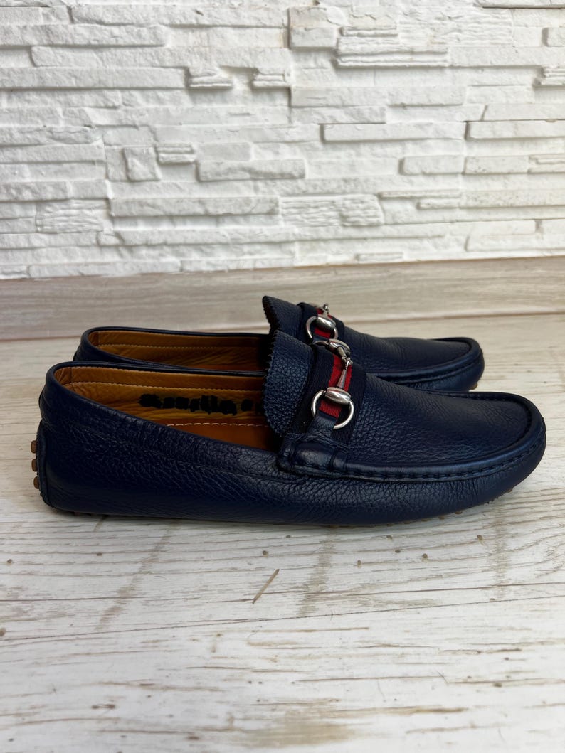 May include: A pair of navy blue leather loafers with a silver-tone metal accent and a red and navy stripe. The shoes are displayed on a light wood surface.
