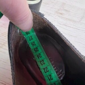 May include: A black shoe with a dark interior lining, shown with a green measuring tape placed inside the footbed. The tape indicates a length measurement extending from approximately 19 to 28 centimeters along the inner sole of the shoe.