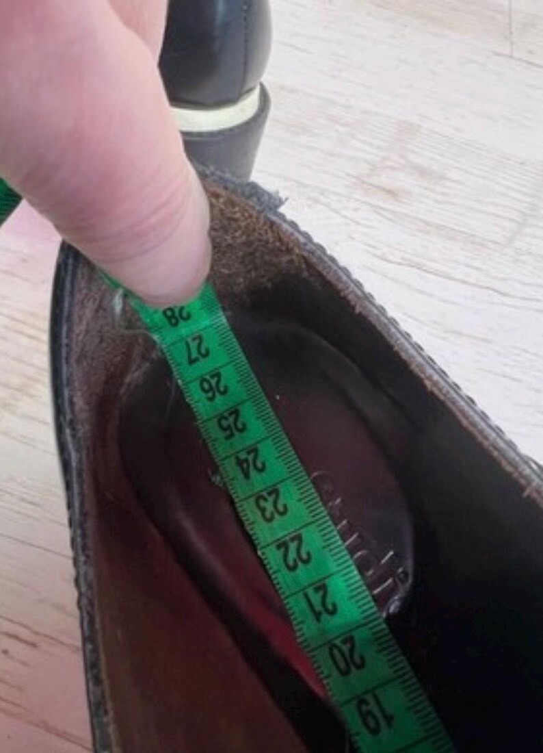 May include: A black shoe with a dark interior lining, shown with a green measuring tape placed inside the footbed. The tape indicates a length measurement extending from approximately 19 to 28 centimeters along the inner sole of the shoe.