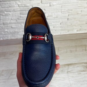 May include: A navy blue leather loafer with a silver-tone metal horsebit detail. The shoe features a red and navy stripe across the vamp. The sole is dark blue. The shoe is held in a hand.