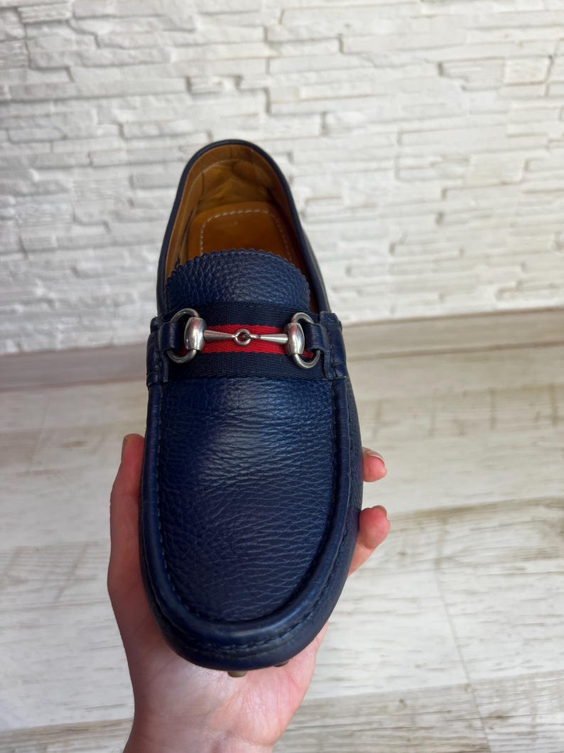 May include: A navy blue leather loafer with a silver-tone metal horsebit detail. The shoe features a red and navy stripe across the vamp. The sole is dark blue. The shoe is held in a hand.
