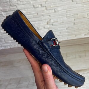 May include: A navy blue leather loafer with a textured finish. The shoe features a silver-tone metal accent with red and green fabric detailing. The sole has small, rounded rubber studs. The shoe is held by a hand.