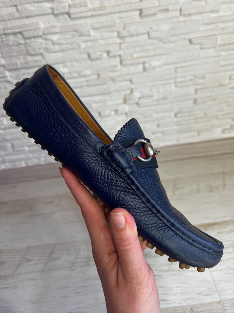 May include: A navy blue leather loafer with a textured finish. The shoe features a silver-tone metal accent with red and green fabric detailing. The sole has small, rounded rubber studs. The shoe is held by a hand.