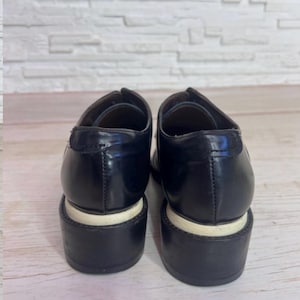 May include: A pair of black leather shoes with a smooth, polished finish and a thick, dark heel. Each shoe features a distinct white horizontal band separating the upper portion from the base of the heel, providing a clear contrast in color.