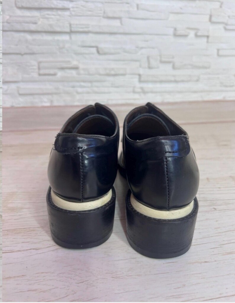 May include: A pair of black leather shoes with a smooth, polished finish and a thick, dark heel. Each shoe features a distinct white horizontal band separating the upper portion from the base of the heel, providing a clear contrast in color.