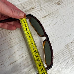 May include: Brown and amber-colored sunglasses with green lenses are measured with a yellow measuring tape. The sunglasses are approximately 6 inches long. The frame has a classic design.