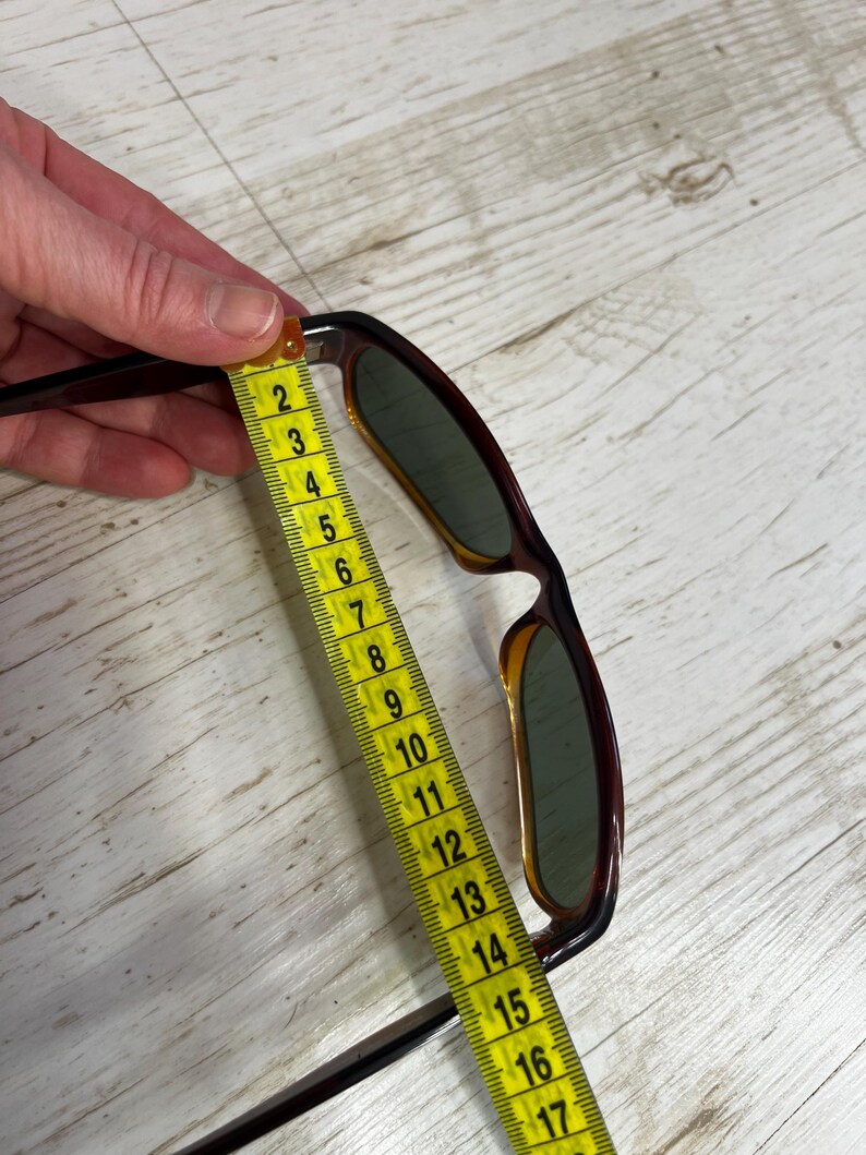 May include: Brown and amber-colored sunglasses with green lenses are measured with a yellow measuring tape. The sunglasses are approximately 6 inches long. The frame has a classic design.
