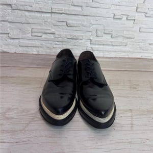 May include: A pair of black leather oxford shoes featuring a pointed toe, black laces, and a thick, contrasting white platform sole. The shoes have a polished, glossy finish and a structured silhouette with a low-cut ankle opening.