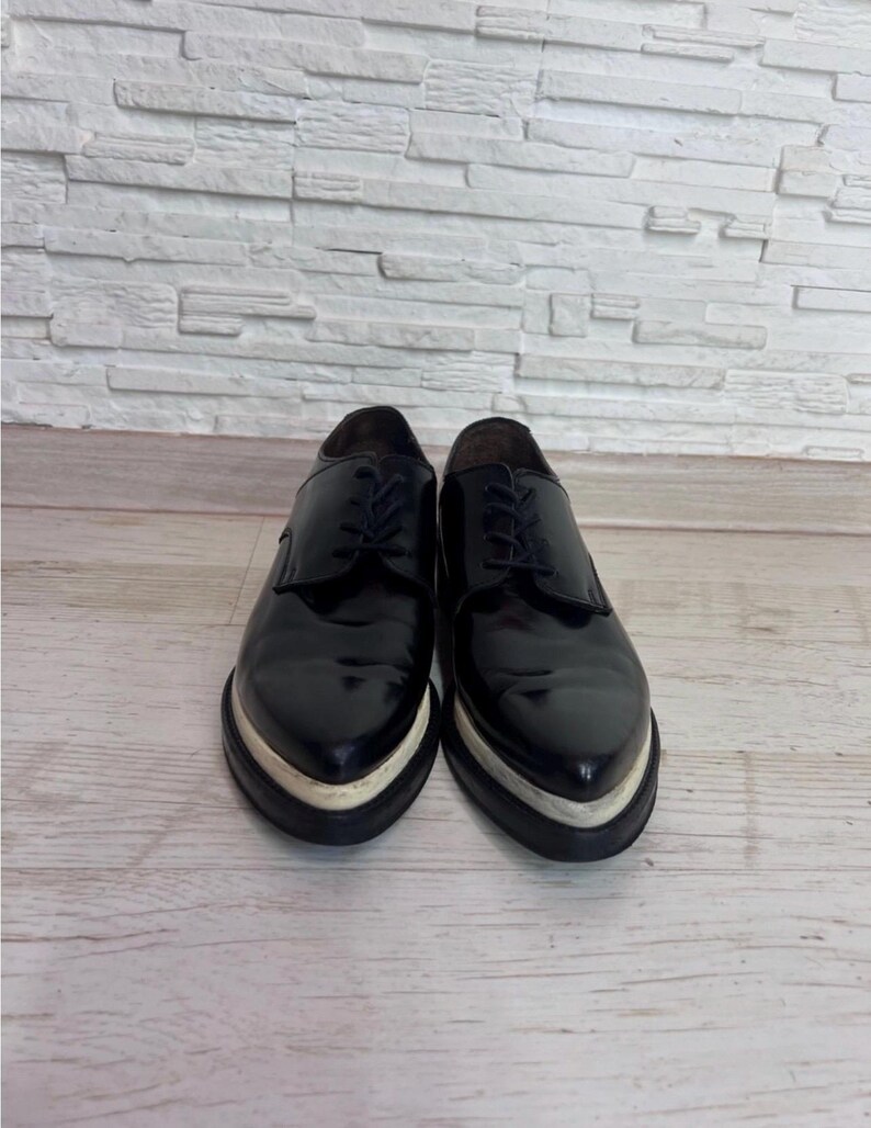 May include: A pair of black leather oxford shoes featuring a pointed toe, black laces, and a thick, contrasting white platform sole. The shoes have a polished, glossy finish and a structured silhouette with a low-cut ankle opening.