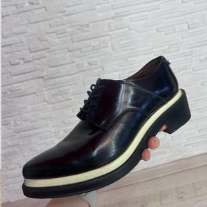 May include: A dark brown leather oxford shoe featuring a lace-up closure, a rounded toe, and a thick, layered platform sole with a distinct cream-colored midsole stripe sandwiched between two black rubber layers, creating a bold profile.