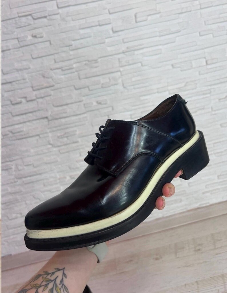 May include: A dark brown leather oxford shoe featuring a lace-up closure, a rounded toe, and a thick, layered platform sole with a distinct cream-colored midsole stripe sandwiched between two black rubber layers, creating a bold profile.