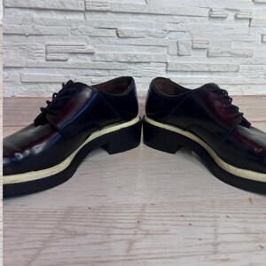 May include: A pair of dark burgundy leather oxford shoes featuring black laces, a rounded toe, and a thick, contrasting white midsole layered above a black rubber outsole. The shoes have a polished finish and a low-cut silhouette with eyelets.