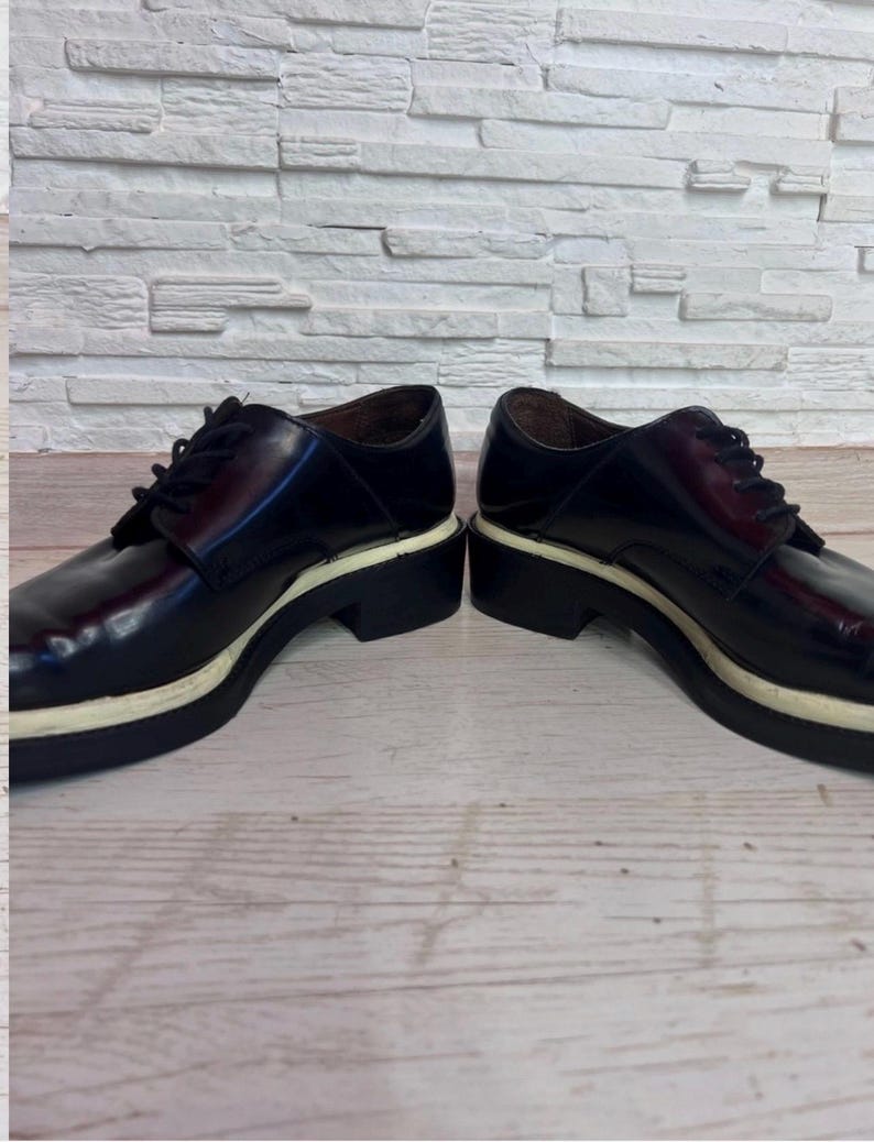 May include: A pair of dark burgundy leather oxford shoes featuring black laces, a rounded toe, and a thick, contrasting white midsole layered above a black rubber outsole. The shoes have a polished finish and a low-cut silhouette with eyelets.