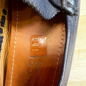 May include: Close-up of a navy blue leather loafer. The shoe has a silver buckle and a stitched upper. The insole is brown leather with the brand name "Gucci" and "made in Italy" printed on it. The shoe is on a light wood surface.