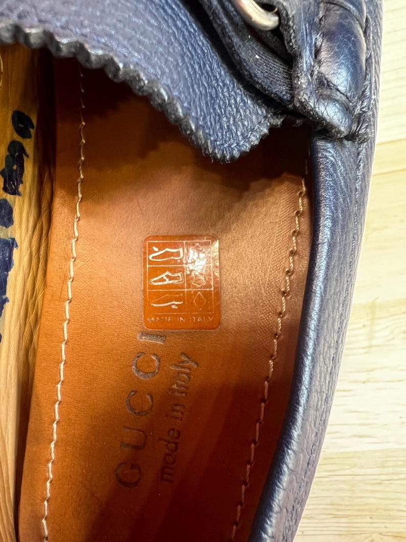 May include: Close-up of a navy blue leather loafer. The shoe has a silver buckle and a stitched upper. The insole is brown leather with the brand name "Gucci" and "made in Italy" printed on it. The shoe is on a light wood surface.