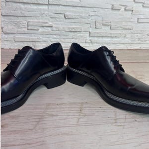May include: A pair of black leather lace-up shoes featuring a smooth, polished finish, black laces, and a thick black sole with a textured, grey patterned welt detail running along the perimeter of the base of the shoe structure.