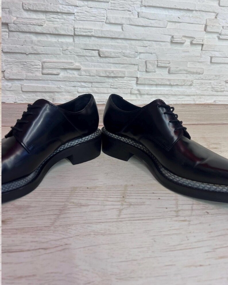 May include: A pair of black leather lace-up shoes featuring a smooth, polished finish, black laces, and a thick black sole with a textured, grey patterned welt detail running along the perimeter of the base of the shoe structure.