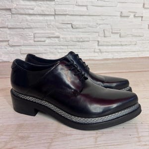 May include: A pair of black leather oxford shoes featuring a lace-up closure, a rounded toe, and a thick black sole with a textured grey snakeskin-patterned trim along the midsole. The shoes have a polished finish and a low block heel.