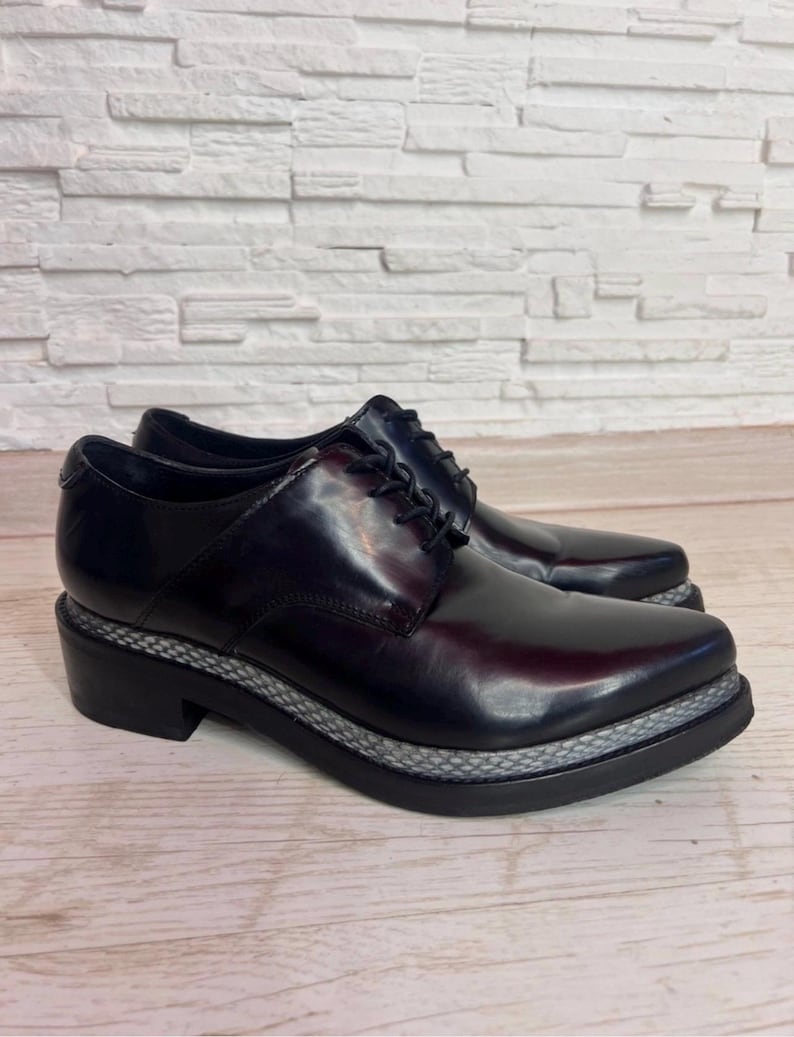May include: A pair of black leather oxford shoes featuring a lace-up closure, a rounded toe, and a thick black sole with a textured grey snakeskin-patterned trim along the midsole. The shoes have a polished finish and a low block heel.