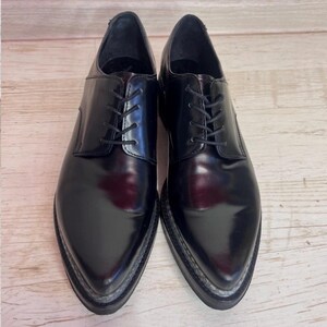 May include: A pair of black leather oxford shoes with a polished finish, pointed toes, and black laces. The shoes feature a thick, dark sole with visible stitching along the edge, providing a structured silhouette for these formal lace-up shoes.