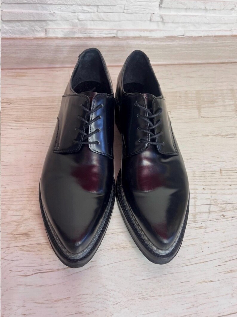 May include: A pair of black leather oxford shoes with a polished finish, pointed toes, and black laces. The shoes feature a thick, dark sole with visible stitching along the edge, providing a structured silhouette for these formal lace-up shoes.