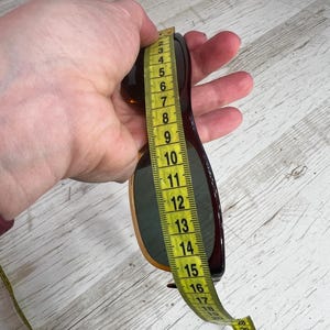 May include: Brown sunglasses with a dark frame are measured with a yellow measuring tape. The sunglasses are held in a hand against a light wood grain background. The sunglasses measure approximately 6 inches in length.