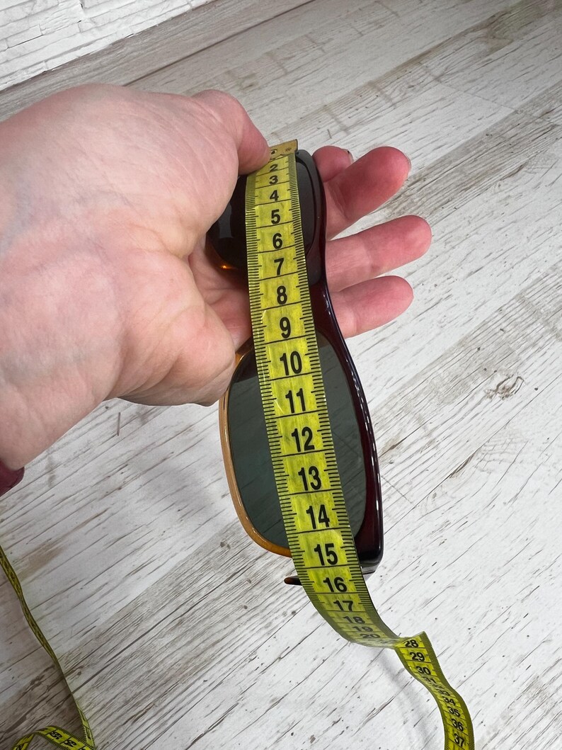 May include: Brown sunglasses with a dark frame are measured with a yellow measuring tape. The sunglasses are held in a hand against a light wood grain background. The sunglasses measure approximately 6 inches in length.