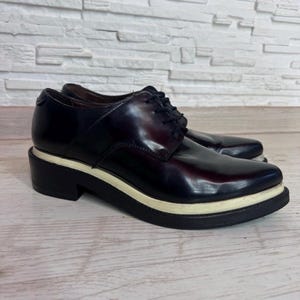 May include: A pair of dark burgundy lace-up oxford shoes featuring a smooth, polished finish, black laces, and a thick, layered sole with a prominent white midsole stripe sandwiched between two black rubber layers for a distinct profile.