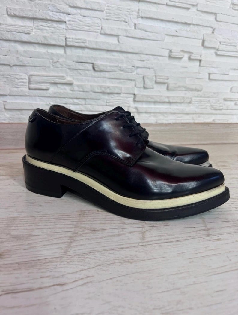 May include: A pair of dark burgundy lace-up oxford shoes featuring a smooth, polished finish, black laces, and a thick, layered sole with a prominent white midsole stripe sandwiched between two black rubber layers for a distinct profile.