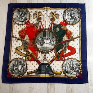 May include: Square silk scarf with a navy blue border and a white centre featuring illustrations of Napoleonic era figures, eagles, and military regalia. The scarf has the word "NAPOLEON" and "HERMES PARIS" printed on it.