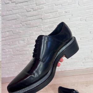 May include: A black lace-up shoe with a smooth, polished finish and a thick, black platform sole. The midsole features a textured, grey and black woven pattern that wraps around the base of the shoe, contrasting with the solid black upper.