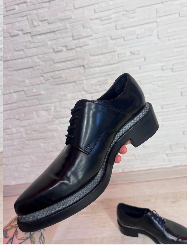 May include: A black lace-up shoe with a smooth, polished finish and a thick, black platform sole. The midsole features a textured, grey and black woven pattern that wraps around the base of the shoe, contrasting with the solid black upper.