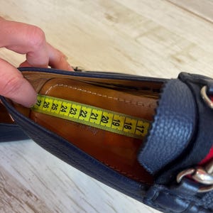May include: Close-up of a navy blue leather loafer with a red and green stripe detail and silver chain accent. A yellow measuring tape shows the shoe's interior length. The shoe appears to be approximately 17 cm long.