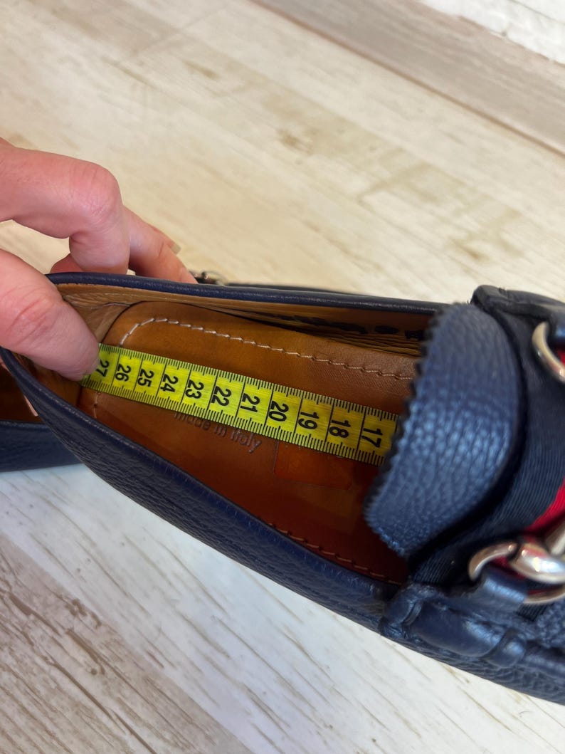 May include: Close-up of a navy blue leather loafer with a red and green stripe detail and silver chain accent. A yellow measuring tape shows the shoe's interior length. The shoe appears to be approximately 17 cm long.