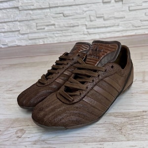May include: A pair of brown leather sneakers with the Adidas logo on the tongue. The shoes have a textured finish and feature the classic three stripes on the sides. The laces and soles are also brown.