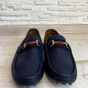 May include: A pair of navy blue leather loafers. Each shoe features a silver-tone metal horsebit detail and a red and navy stripe across the top. The soles are a light brown color.
