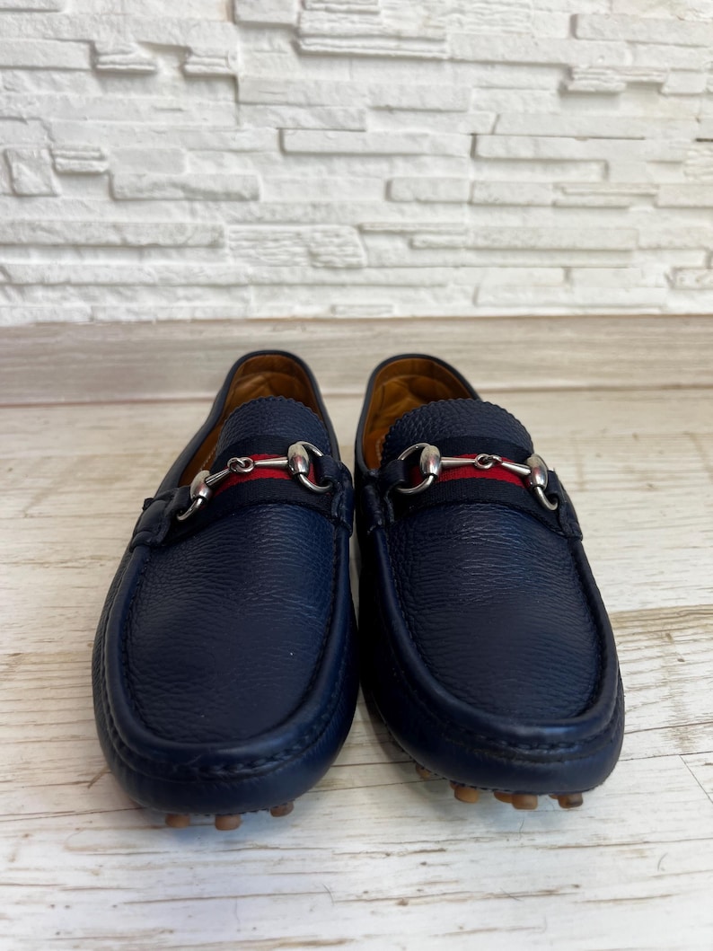 May include: A pair of navy blue leather loafers. Each shoe features a silver-tone metal horsebit detail and a red and navy stripe across the top. The soles are a light brown color.