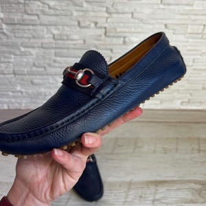 May include: Navy blue leather loafer with a silver-tone metal horsebit detail. The shoe has a textured leather upper and a rubber sole. The loafer is a classic style, suitable for casual or semi-formal wear.
