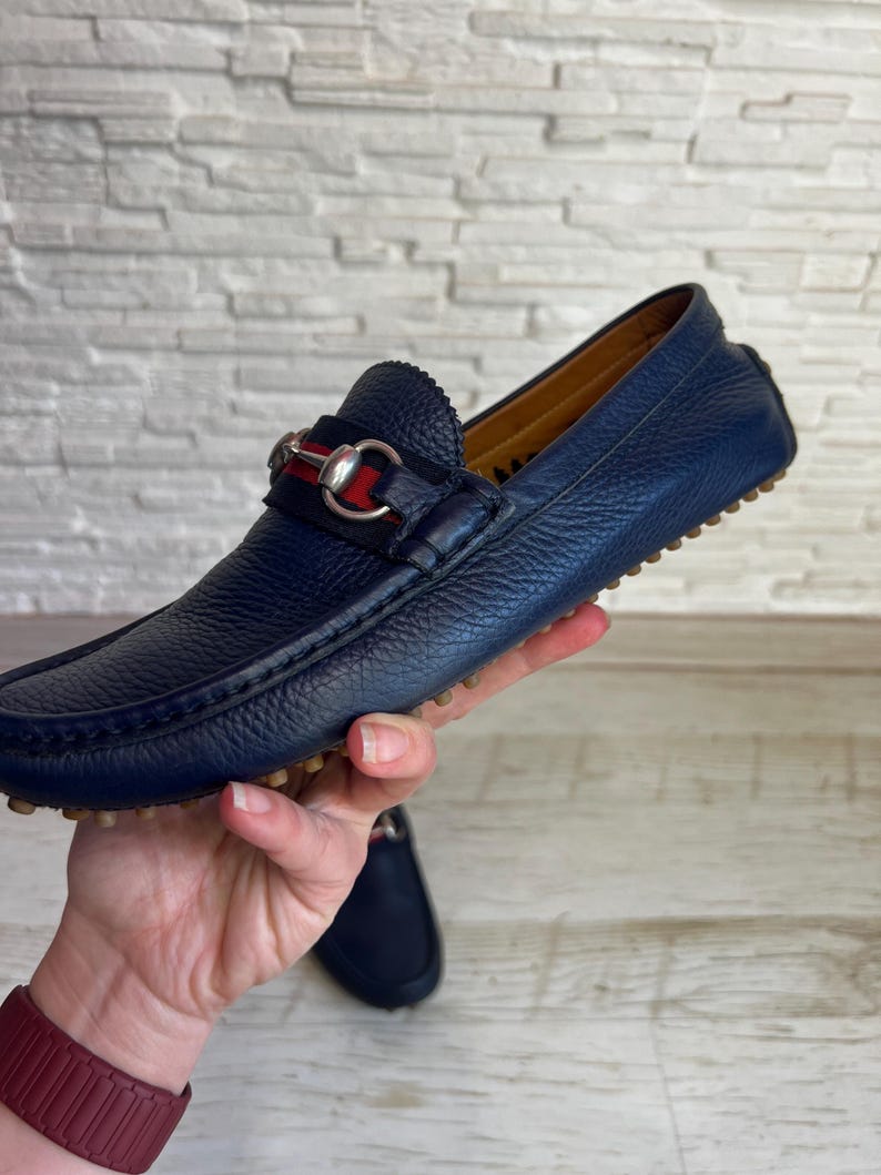 May include: Navy blue leather loafer with a silver-tone metal horsebit detail. The shoe has a textured leather upper and a rubber sole. The loafer is a classic style, suitable for casual or semi-formal wear.