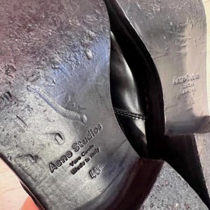 May include: The black leather soles of a pair of shoes featuring the embossed text Acne Studios, Vero Cuoio, Made in Italy, and the size 40. The soles show signs of wear with a smooth finish and a stacked heel, displaying clear branding details.