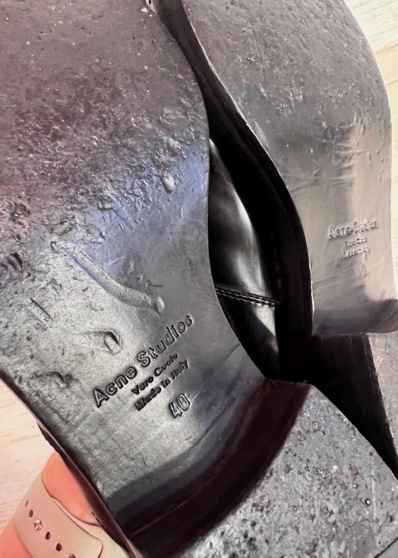 May include: The black leather soles of a pair of shoes featuring the embossed text Acne Studios, Vero Cuoio, Made in Italy, and the size 40. The soles show signs of wear with a smooth finish and a stacked heel, displaying clear branding details.