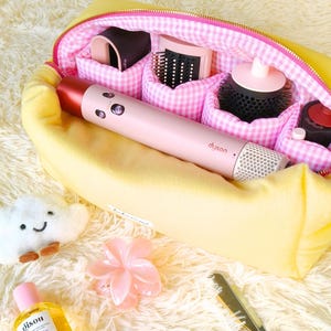 May include: A yellow zippered pouch with a pink gingham interior containing a pink Dyson hair styling tool and attachments. Outside are a white cloud plush, a pink flower hair clip, a bottle of Gisou hair oil, two Rhode lip tints, and a gold comb.