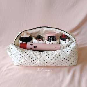 May include: A white quilted storage bag with a black polka dot pattern containing a pink Dyson hair styling tool and several cylindrical hair styling attachments. The bag features a black zipper closure and the text kokoromi printed below it.