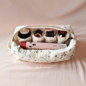 May include: A quilted cream-colored travel case with a small floral pattern containing a pink Dyson hair styling tool and four cylindrical attachments. The case features a gold-toned zipper and internal dividers to organize the hair styling components.