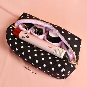 May include: A black fabric travel case with white polka dots and a gold zipper, containing a light pink Dyson hair styling tool and several round brush attachments. The interior is lined with a light pink and white striped fabric.