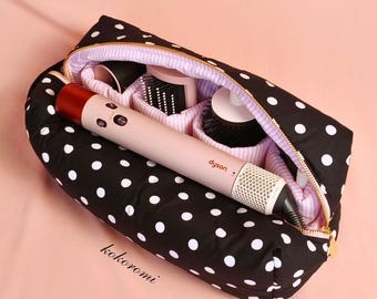 May include: A black fabric travel case with white polka dots and a gold zip, containing a light pink Dyson hair styling tool and several round brush attachments. The interior is lined with a light pink and white striped fabric.