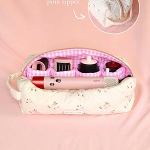 May include: A quilted cream-coloured travel case with a small pink floral pattern, featuring a pink and white gingham interior lining with four padded compartments. A pink Dyson hair styling tool is placed horizontally inside the open bag.