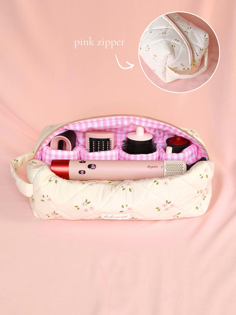 May include: A quilted cream-coloured travel case with a small pink floral pattern, featuring a pink and white gingham interior lining with four padded compartments. A pink Dyson hair styling tool is placed horizontally inside the open bag.