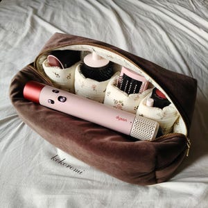 May include: A brown velvet zippered storage bag containing a light pink Dyson hair styling tool and several hair styling attachments nestled in individual cream-colored fabric compartments with a delicate floral pattern and the text kokoromi.